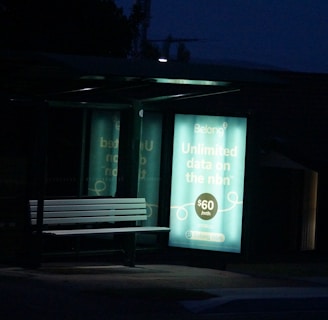 A dimly lit bus stop bench is illuminated by an advertisement panel displaying an internet service promotion. The surrounding area is dark, with the advertisement text prominently visible.