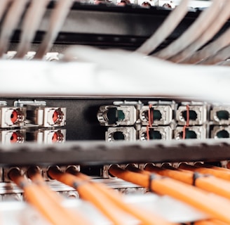 A close-up view of a network server or data center with numerous cables and connections. Orange cables are neatly arranged in the foreground, while various ports and connectors are visible in the background.