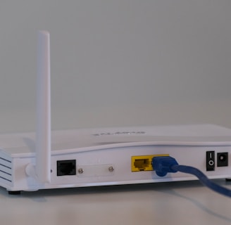 A white wireless router with two antennas is placed on a flat surface. The back of the router is visible, showing various ports including a yellow Ethernet port with a blue cable connected, power input, and a power switch.